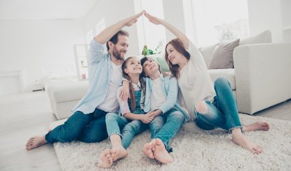 New building residential house purchase apartment concept. Stylish full family with two kids sitting on carpet, mom and dad making roof figure with hands arms over heads