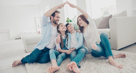 New building residential house purchase apartment concept. Stylish full family with two kids sitting on carpet, mom and dad making roof figure with hands arms over heads